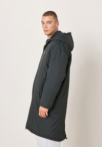 Muž stojaci bokom proti jednoduchému svetlému pozadiu, oblečený v dlhom čiernom prešívanom kabáte s kapucňou.