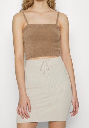 Vrouw draagt een pluizige bruine cropped tanktop en een beige geribbelde rok met hoge taille en een strikdetail aan de voorkant, staand tegen een witte achtergrond.