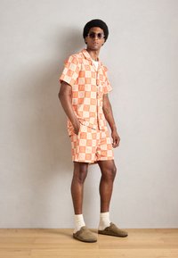 Rövid ujjú, narancssárga mintás ing és bermuda szett, könnyű anyagból készült. Fehér geometrikus mintákkal díszítve, barna cipővel párosítva.