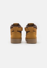 Une paire de baskets montantes marron et beige, tournées vers l'arrière, avec des cols rembourrés et des sangles réglables, sur un fond blanc.