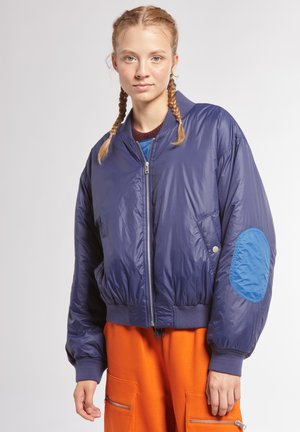 Giacca bomber blu navy con zip frontale, polsini a costine e una toppa decorativa sul gomito. Materiale liscio e lucido con una consistenza leggermente imbottita.