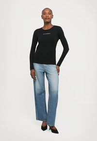 Vrouw die staat in een aansluitend zwart longsleeve shirt met witte tekst, lichtblauwe wijde jeans en zwarte puntige hakken.