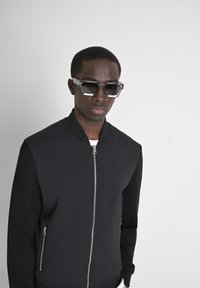Giubbotto bomber nero con una parte anteriore testurizzata, maniche lisce e tasche con zip; abbinato a occhiali da sole angolari bicolore.