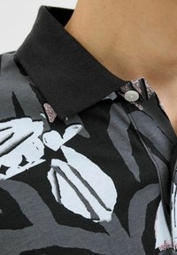 Polo tričko s límečkem v vzorované černé, šedé a světle modré látce. Má černý límeček, zapínání na knoflíky a jemný květinový design.