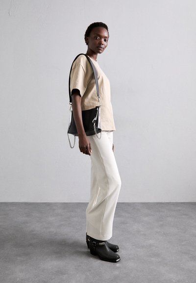 Beige kortærmet top, hvide lige ben bukser og sorte hælede klipklapper. Sort håndtaske med kæde detaljer på remmen. Neutral baggrund.
