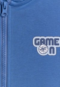 Tissu bleu avec une fermeture éclair sur la gauche et texte blanc « GAME ON » avec un graphisme de volley-ball et logo « 4F » sur le côté droit.