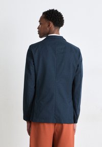 Blazer bleumarin, ajustat, fabricat dintr-un material texturat, cu guler perforat și design cu un singur nasture, asortat cu pantaloni scurți portocalii.