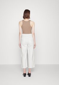 Canottiera marrone e pantaloni bianchi a gamba larga, con orlo grezzo. Design aderente con una texture liscia, abbinati a scarpe con tacco quadrato.