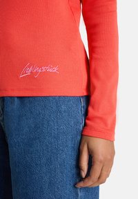 Person in einem langärmligen, gerippten korallfarbenen Oberteil mit "Liedingsstick" auf Höhe des Saums gestickt, und blauen Denim-Jeans, die Hand entspannt an der Seite.