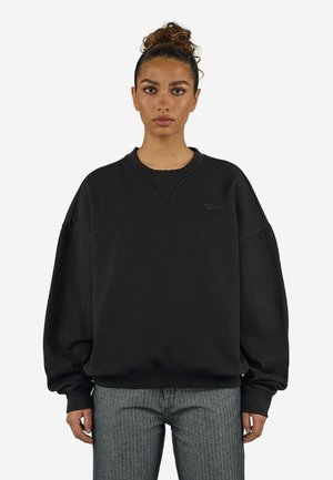 Ung kvinde med hår i en knold iført en oversized sort sweatshirt og grå jeans, stående mod en ensfarvet lys baggrund.