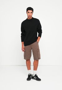 Pull en maille noir avec des poignets côtelés, associé à un short cargo marron clair avec des poches latérales. Baskets noires avec des accents blancs.