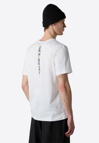 Hvid crew-neck t-shirt med korte ærmer, der har et vertikalt sort og gråt logo-print på ryggen. Båret med en sort strikket hue.