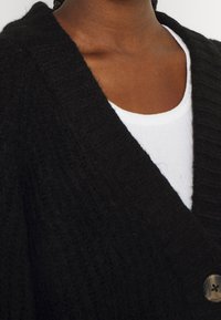 Dark-skinned person wearing a black textured cardigan over a white ribbed crewneck shirt, button visible on the cardigan.