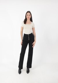 Top ajustado beige con escote cuadrado, combinado con pantalones negros de cintura alta que cuentan con botones dorados y un diseño recto. Botas negras de tacón.