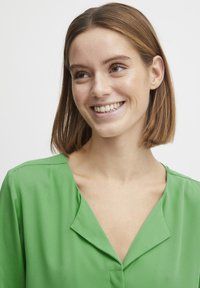 Green, lightweight blouse with a V-neck and subtle fold detailing. Smooth texture with a relaxed fit and short sleeves.