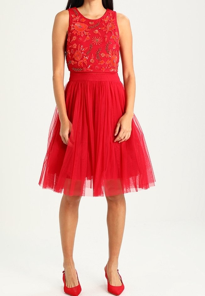 Femme portant une robe rouge sans manches avec une broderie florale sur le corsage et une jupe en tulle, associée à des talons hauts rouges.