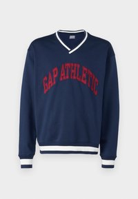 Granatowa bluza z dekoltem w szpic, z napisem "GAP ATHLETIC" w czerwonym, łukowatym kształcie. Prążkowane, białe i granatowe mankiety oraz dół. Miękka faktura materiału.