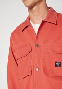 Man som bär en röd knäppjacka med fickor och en Timberland-logga på bröstet över en vit skjorta.