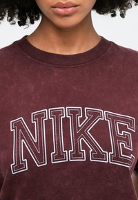 Burgundijas kokvilnas T-krekls ar lielu baltu kontūra "NIKE" logotipu. Audums ir mīksts ar nedaudz teksturētu virsmu.