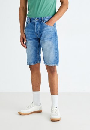 Mann in hellblauen Jeansshorts, weißen Socken, weißen Sneakern und einem grünen Shirt, der vor einem schlichten weißen Hintergrund steht.