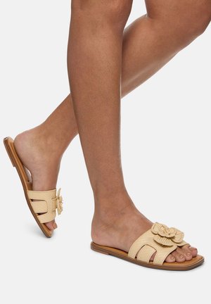 Sandali open-toe beige con una finitura testurizzata. Presentano un accento floreale sulla cinghia e una suola piatta e flessibile con un leggero arco.