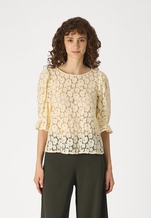 Lace bluse i lys gul med blomstermønster, rund halslinje og puffete ermer. Hempeket går over i mørkere blonder nederst.