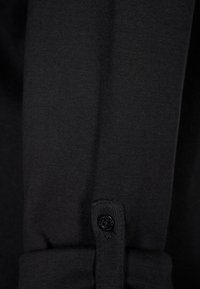 Détail d'une chemise noire à manches longues avec une texture de tissu lisse et un poignet avec un seul bouton noir.