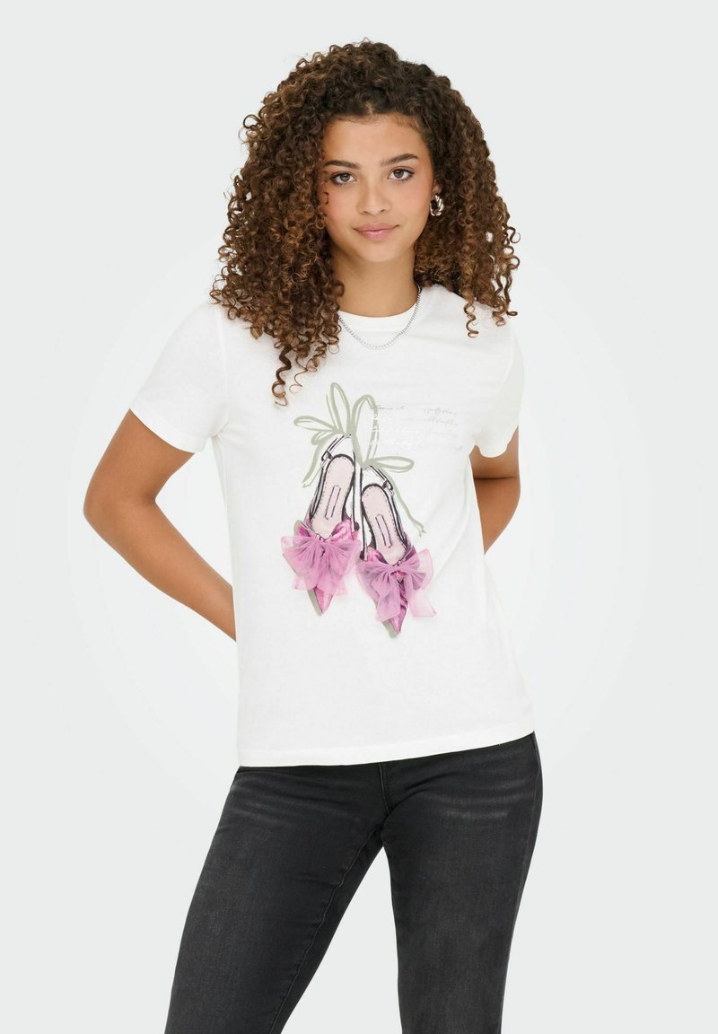 Jonge vrouw met krullend haar, gekleed in een witte t-shirt met een afbeelding van roze balletschoenen met linten en zwarte jeans, staande met haar handen achter haar rug.