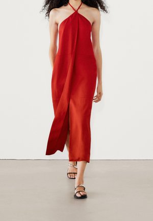 Femme marchant dans une robe midi rouge sans manches à col halter avec une fente à l'avant, associée à des sandales à talons noires à brides.