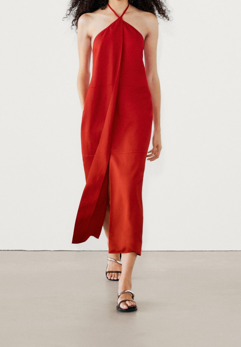 Femme marchant dans une robe midi rouge sans manches à col halter avec une fente à l'avant, associée à des sandales à talons noires à brides.