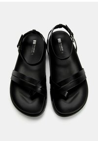 Sandales noires en matériau synthétique lisse, avec des brides croisées et une sangle de cheville ajustable. Semelle plate et design minimaliste.