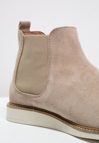 Béžová semišová Chelsea obuv s elastickým bočním panelem, poutko na zadní straně a bílou gumovou podrážkou s hnědým prošívaným okrajem.