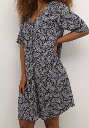 Kvinde iført en kortærmet kjole med V-hals, sort bundfarve og flerfarvet paisley- og blomsterprint, hånd hvilende på hoften og sidelomme.