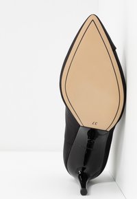 Chaussure à talon haut noire avec un bout pointu et un talon fin. Le matériau est lisse avec une finition mate, comprenant une semelle beige et un contour subtil. Taille 37.