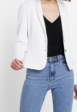 Person, der bærer en tekstureret hvid blazer over en sort V-hals top og højtaljede blå jeans mod en ensfarvet baggrund.