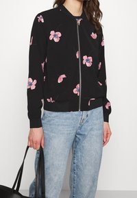 Svart bomberjacka med rosa blommönster, dragkedjestängning, ribbade mudd och nederkant, lättviktig tyg, kombinerad med ljusblå jeans.