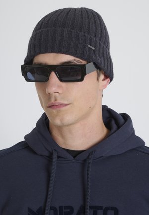 Donkergrijze geribde gebreide muts gecombineerd met oversized zwarte rechthoekige zonnebril. Model draagt een marineblauwe hoodie met een trekkoordhals.