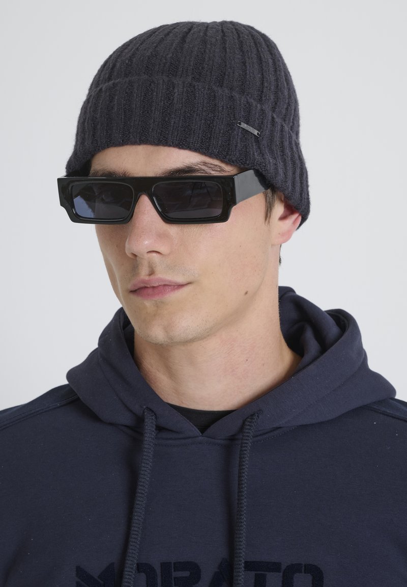 Donkergrijze geribde gebreide muts gecombineerd met oversized zwarte rechthoekige zonnebril. Model draagt een marineblauwe hoodie met een trekkoordhals.