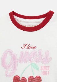 Weißes T-Shirt mit einem roten Rippkragen. Auf der Vorderseite ist "I love Guess" sowie Kirschen in Pink, Rot und Grün mit dem Retro-Jahr "1981" aufgestickt.