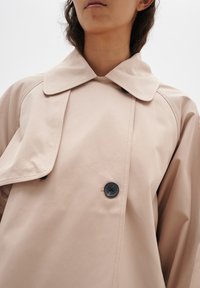 Beige trench coat, jossa on leveä kaulus, kaksirivinen malli mustilla napilla ja sileä, kevyt kankaan rakenne.