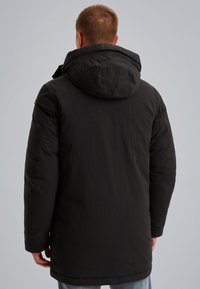 Zwarte gewatteerde jas met een capuchon, met een gladde textuur en een langere snit. Bevat een subtiele glans en minimale hardware-accenten.