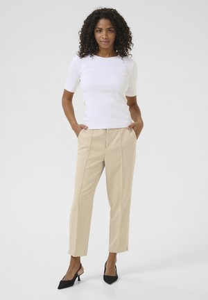 Haut blanc côtelé à manches courtes associé à un pantalon beige ajusté et des escarpins noirs à bride arrière ; le modèle se tient les mains dans les poches, sur un fond neutre.