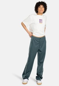 White t-shirt with graphic text, paired with teal athletic pants featuring white side stripes, worn with white sneakers.