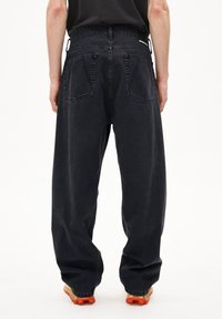 Pantalons en denim noir à coupe ample, taille haute, avec deux poches arrière. Des coutures distinctives et une finition texturée contrastent avec le tissu lisse.