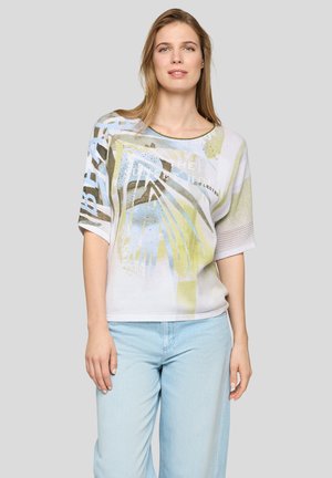 Frau trägt einen hell gemusterten, lockeren Pullover mit pastellblauem, grünem und beigem abstraktem Design, kombiniert mit hellblauen Jeans.
