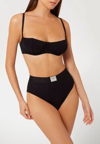 Crni rebrasti bikini set koji se sastoji od strukturirane gornje strane s žicom i tankim trakama, te visokih gaćica s pojasom sa brandom.