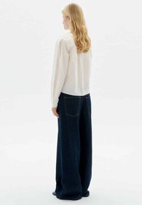 White long-sleeve blouse with a buttoned back, paired with loose-fitting dark denim jeans featuring two back pockets and contrasting stitching.