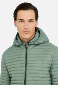 Hættepuffjakke i dæmpet grøn med horisontal quiltet tekstur, en frontlynlås og justerbar hætte med elastisk kant.