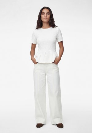 Femme debout les mains dans les poches portant un haut péplum blanc texturé à manches courtes, un pantalon blanc à jambes larges et des chaussures marron foncé sur fond blanc.