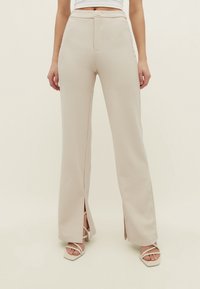 Pantalons larges beige avec fentes avant, tissu texturé, taille haute et fermeture à bouton, associés à des talons fins de couleur claire.